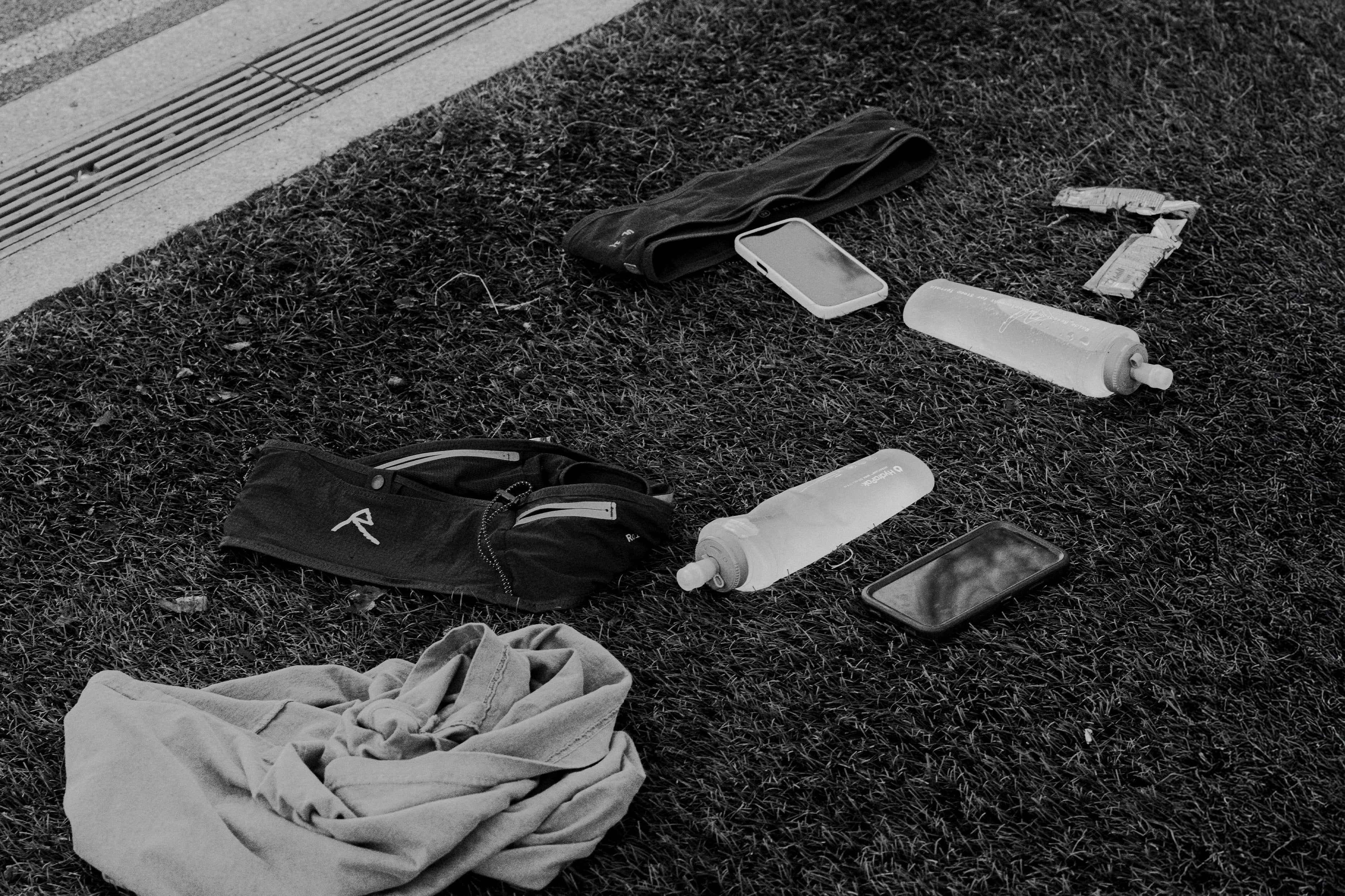 A black and white image of trail running gear resting on grass, featuring a Raide Research pack, two Hydrapak soft flasks, smartphones, a gel wrapper, and apparel.