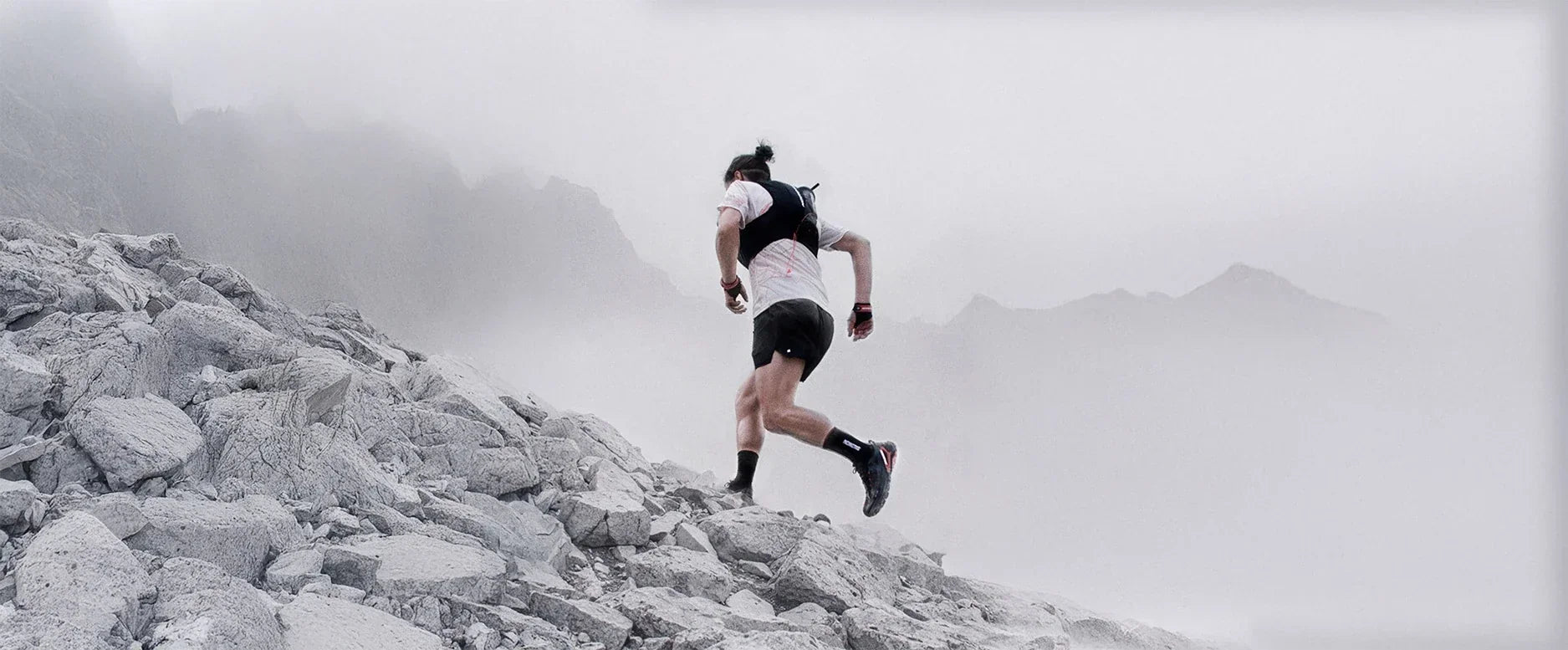 A trail runner wearing a Salomon hydration vest ascends a steep, rocky mountain slope in heavy fog.