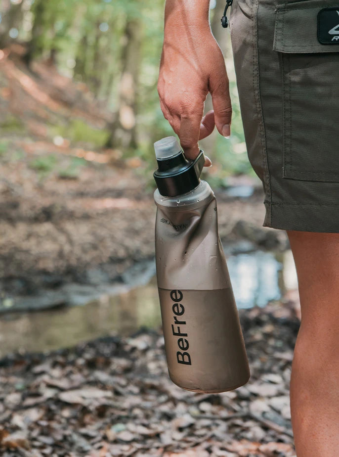 Close-up of a hiker holding a tactical tan Katadyn BeFree water filter flask by its cap handle while walking through a forest.