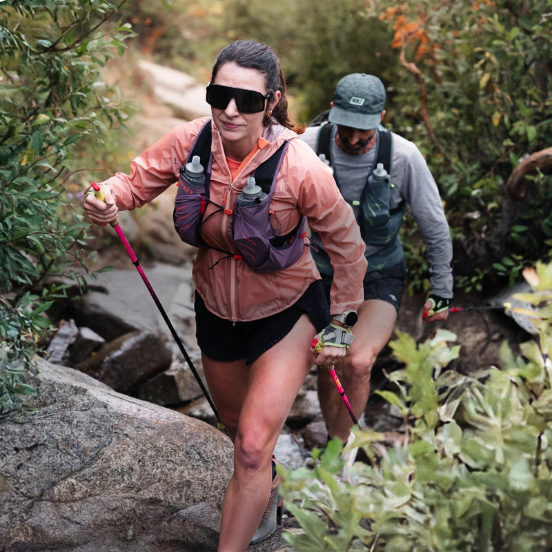 Two trail runners navigating a rocky path through brush, wearing Osprey hydration vests equipped with front soft flasks.