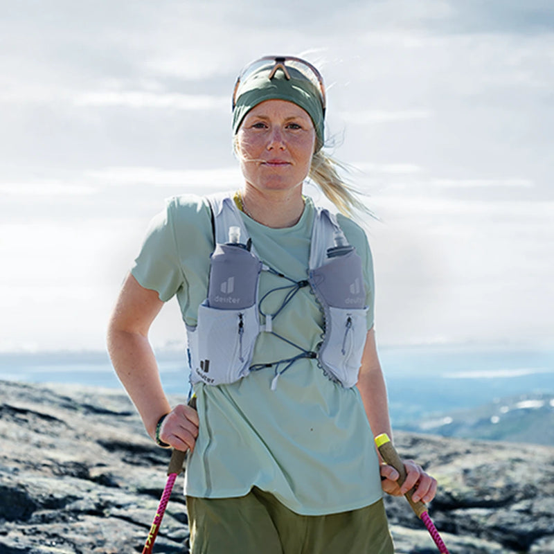 A female athlete standing on a rocky mountain expanse holding trekking poles, wearing a deuter hydration vest with two front soft flasks.