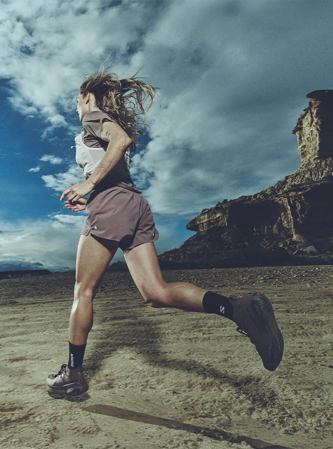 A side view of a female runner striding across a rocky, desert-like plateau under a dramatic cloudy sky, wearing a light grey Salomon running vest.