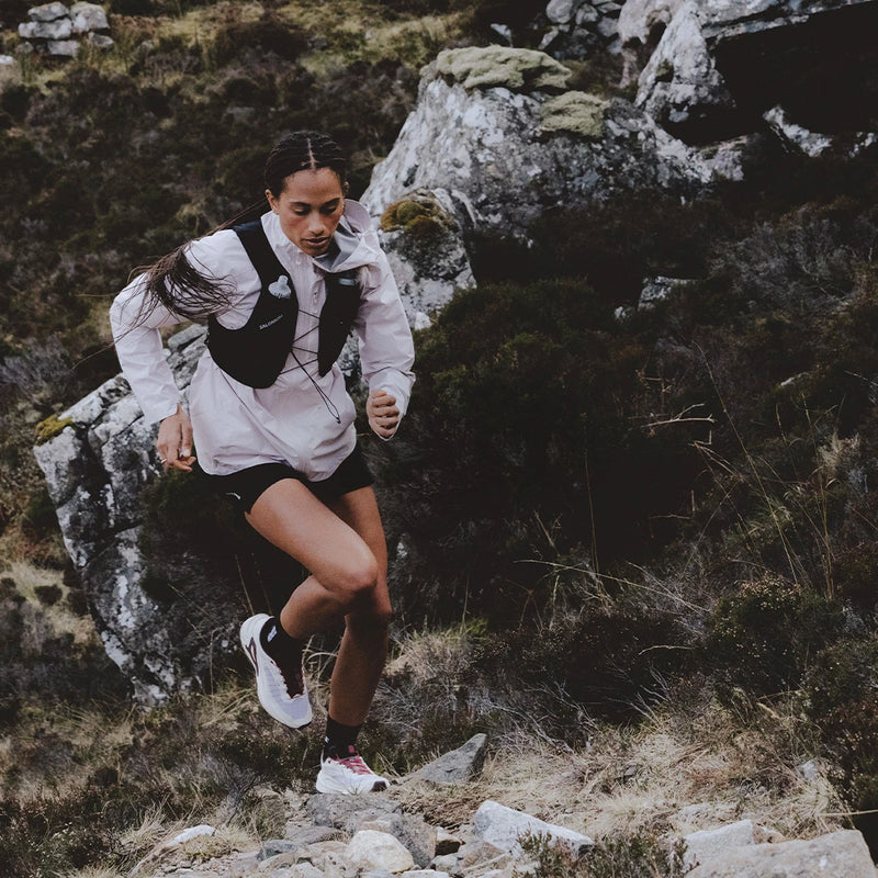 A female runner sprinting along a rugged, rocky mountain trail wearing a white jacket and a black Salomon running vest.