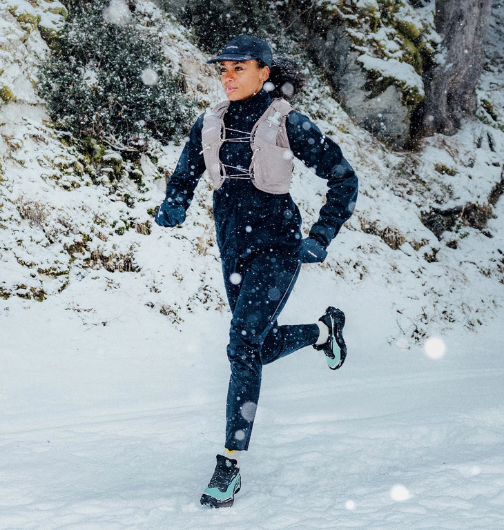 A female athlete trail running through snowy terrain during snowfall, wearing a light grey Salomon running vest over dark cold-weather apparel.