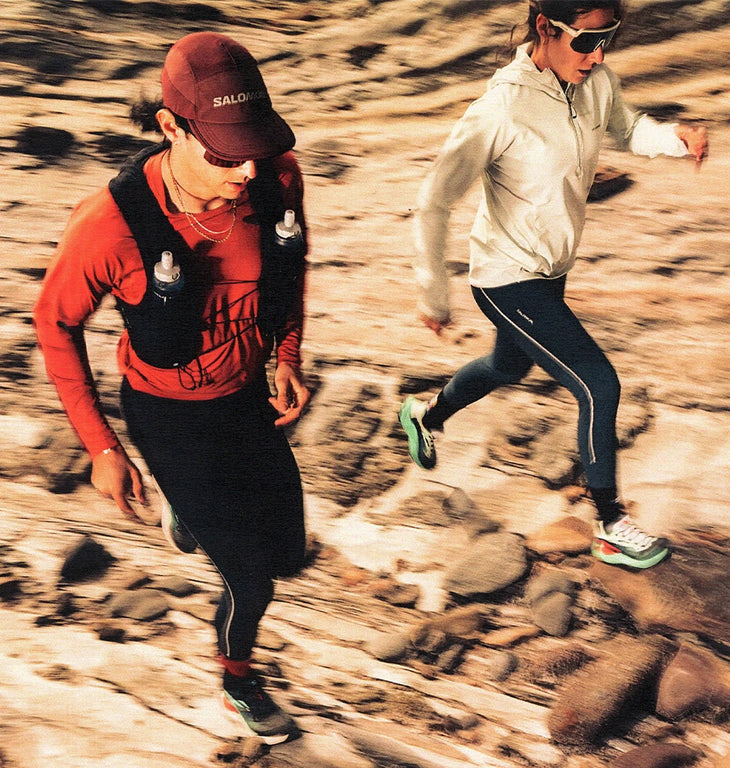 Two athletes running on a technical dirt trail, highlighting a Salomon S/LAB hydration vest with HydraPak soft flasks held in front chest pockets.