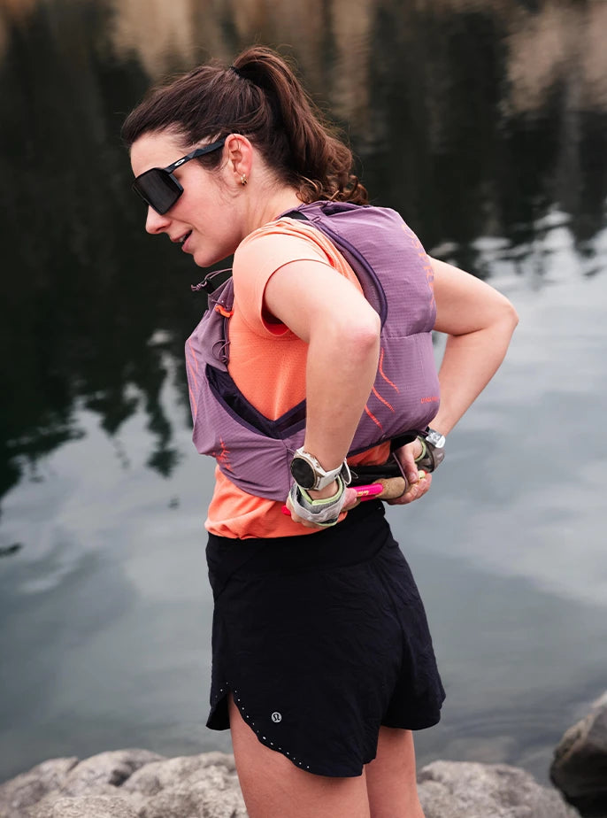 A female trail runner standing by a lake, reaching behind her to stow her trekking poles on the back of her purple Osprey hydration vest.