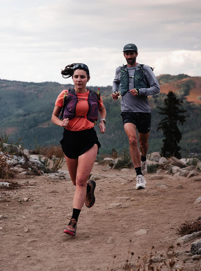 A female and male athlete running on a dirt trail, equipped with Osprey hydration packs and soft flasks.