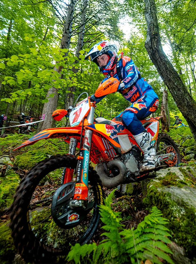 A dirt bike athlete wearing a USWE hydration pack with a red drink tube, riding through a forested enduro course.