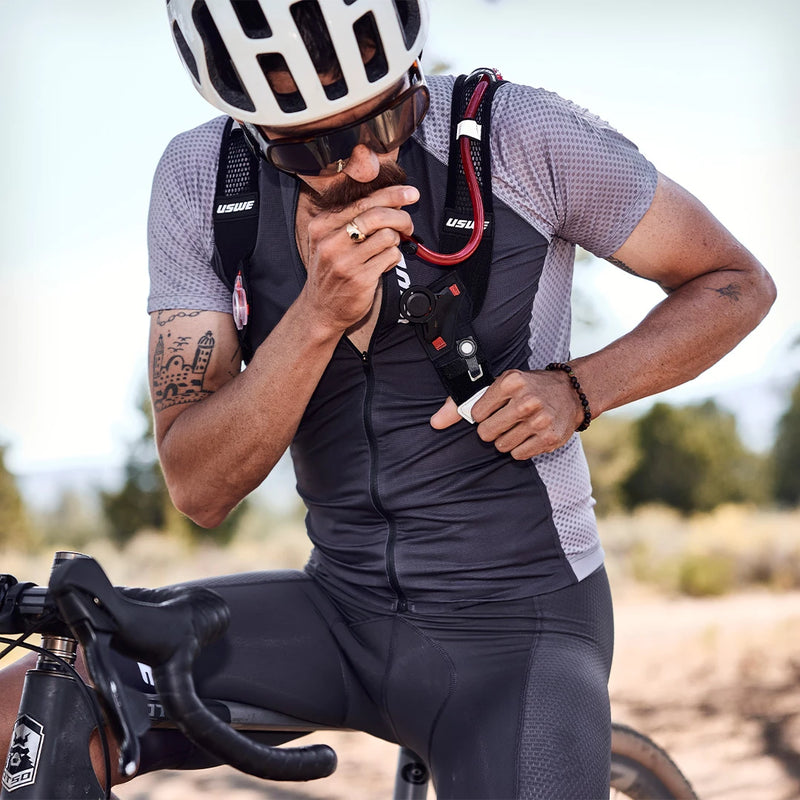 Close-up of a rider on a bicycle drinking via USWE’s signature red HydraPak hydration tube while adjusting his harness strap.